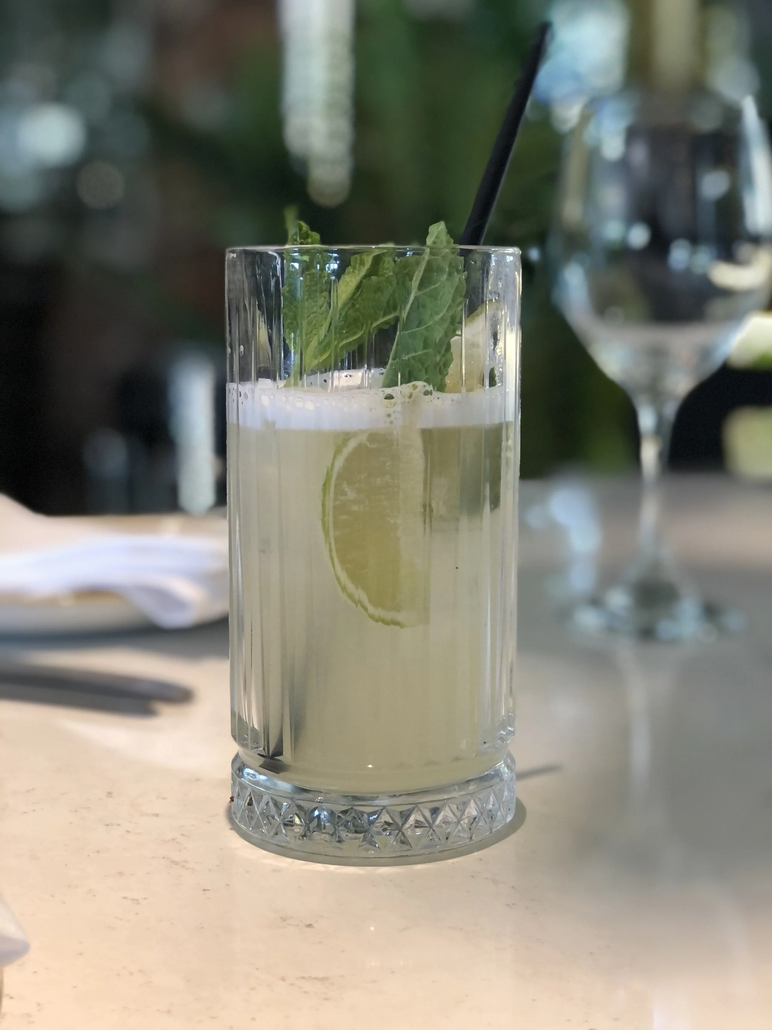 A tall glass of lemonade with lemon slice and mint leaves, with a black straw inside, on a white table near a wine glass.