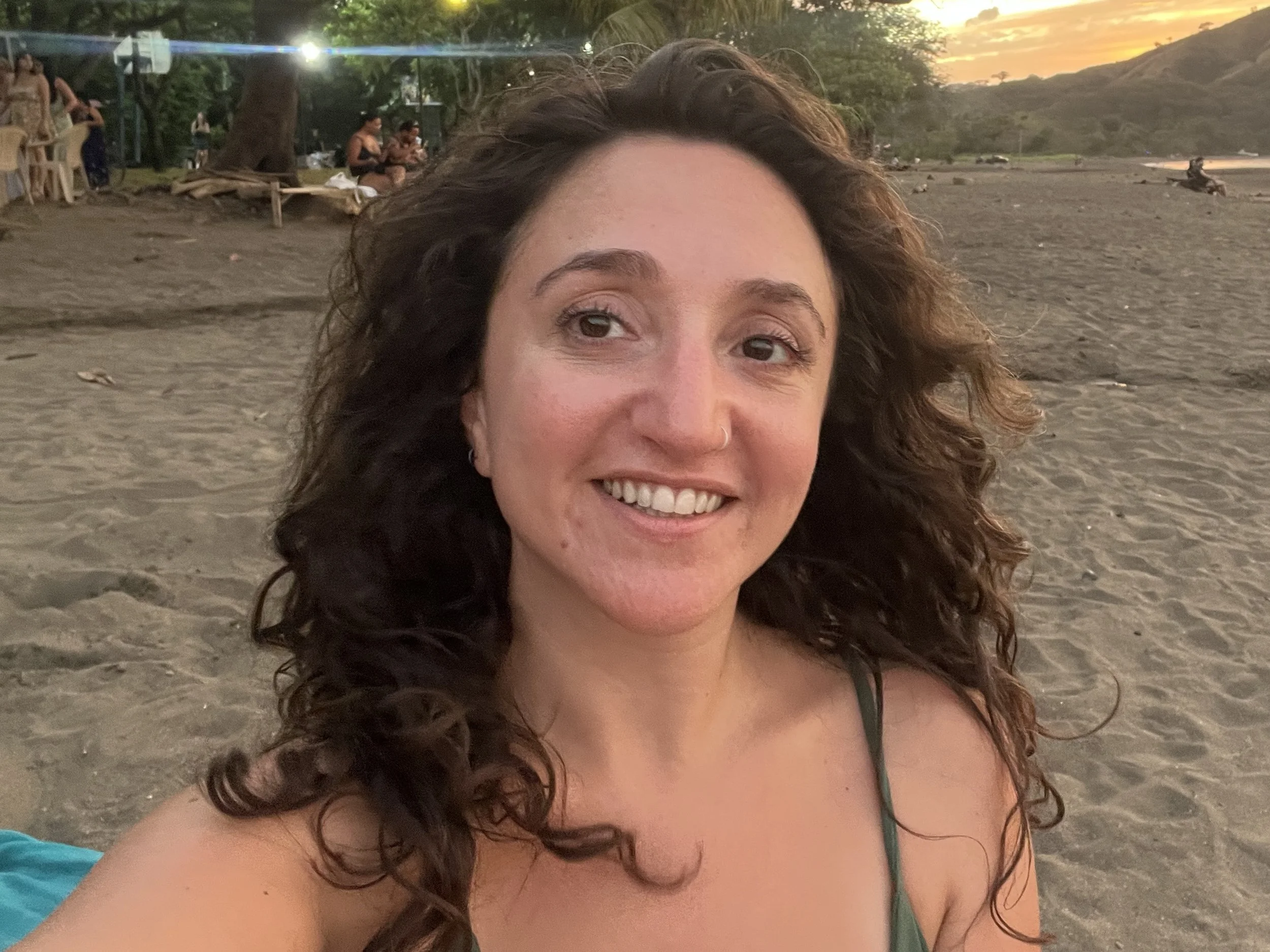 A woman with curly brown hair smiling at the beach during sunset, with a sandy shoreline and hills in the background.