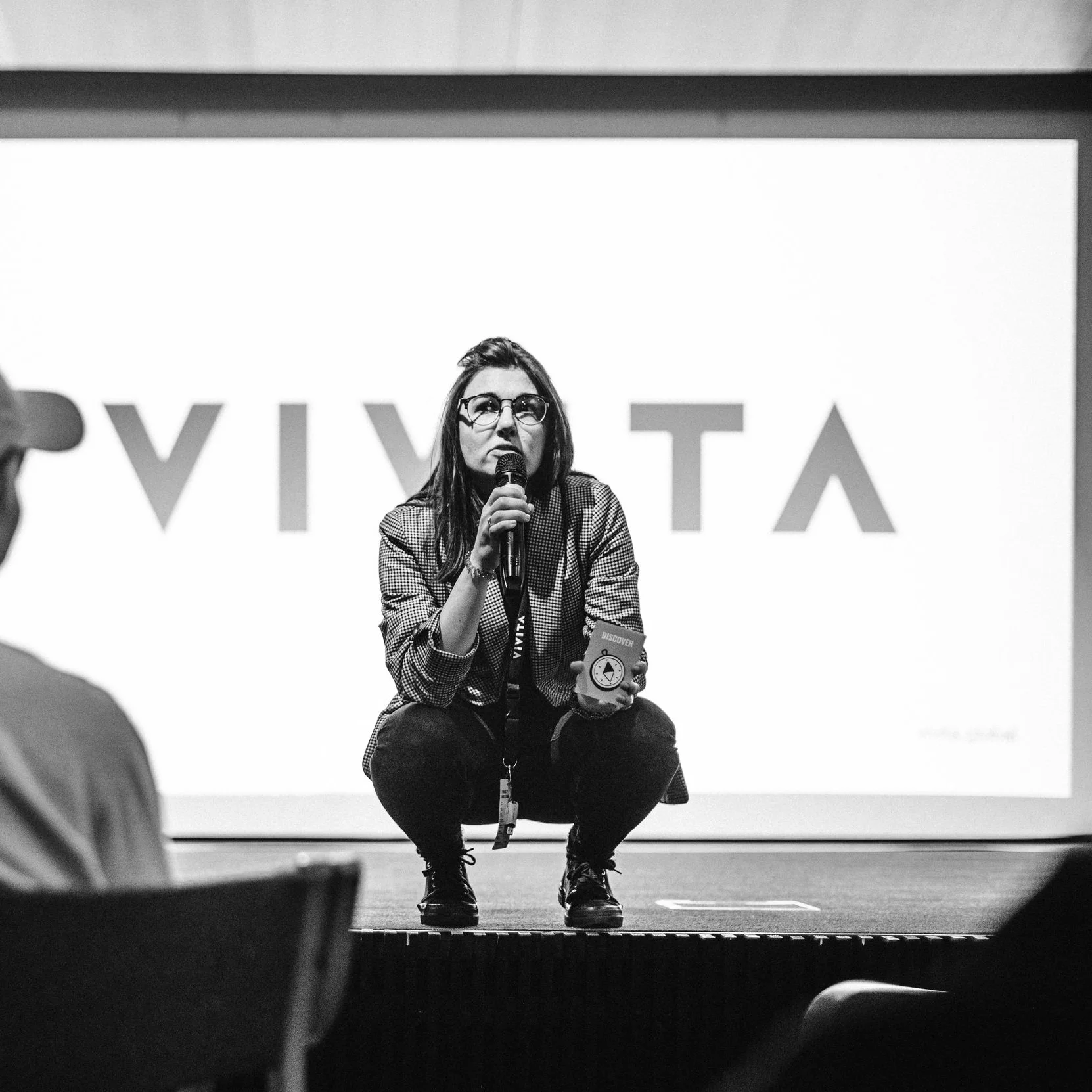 Person crouching on stage holding a microphone and a small object, with a large screen displaying partial text in the background.