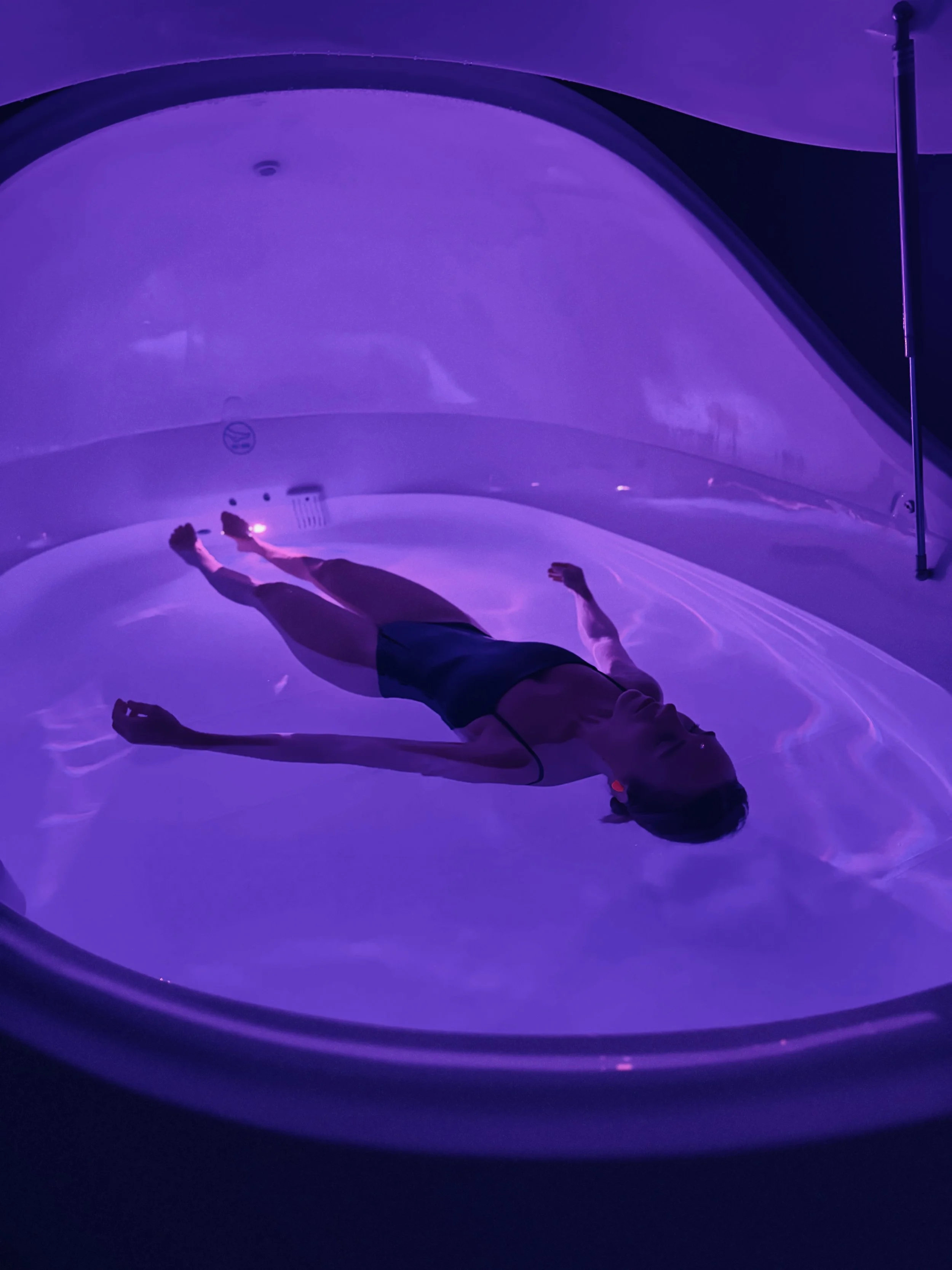 Person lying on a purple-lit table or pod, with their arms stretched out and legs extended, inside a dark, futuristic-looking chamber.