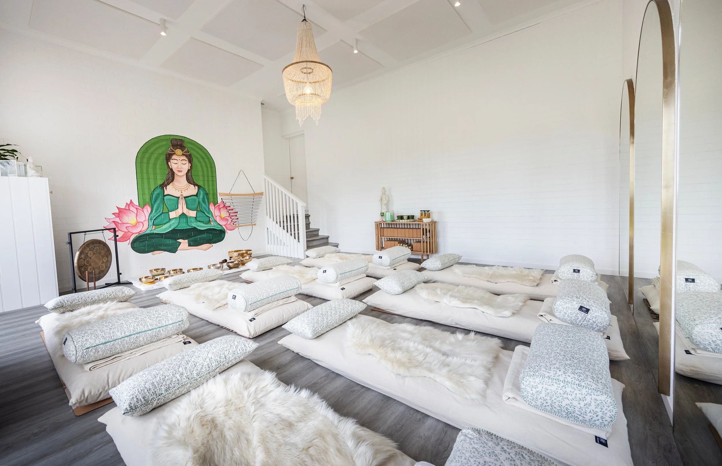 A peaceful meditation space with a large mural of a woman in a prayer pose, surrounded by pink lotus flowers on the wall. The room has white walls and ceiling, a chandelier, and floor cushions arranged on a soft white rug for sitting or meditation.