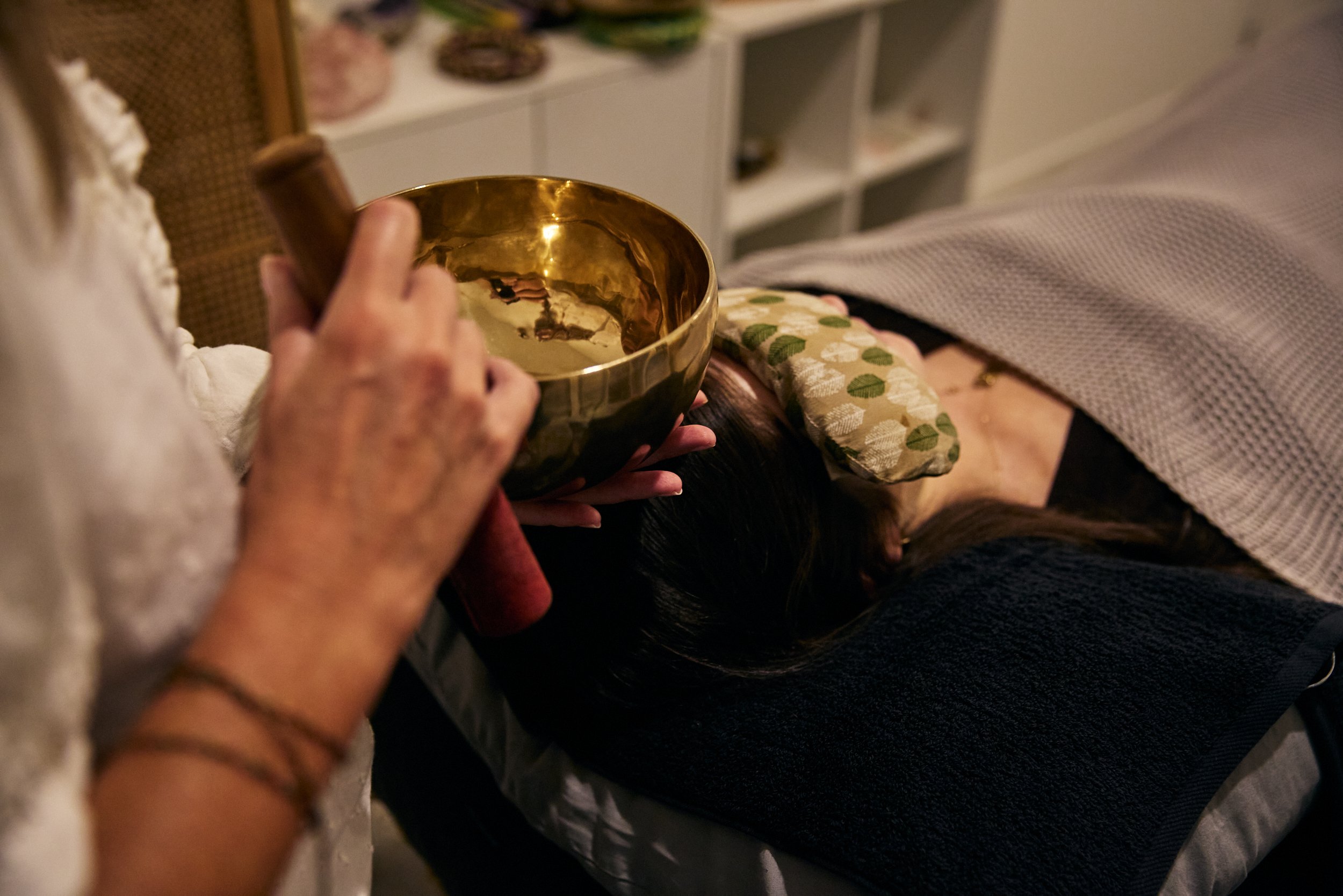 An elderly woman holding a singing bowl near a person's head who is lying down, possibly receiving a sound therapy or meditation session, in a cozy room.