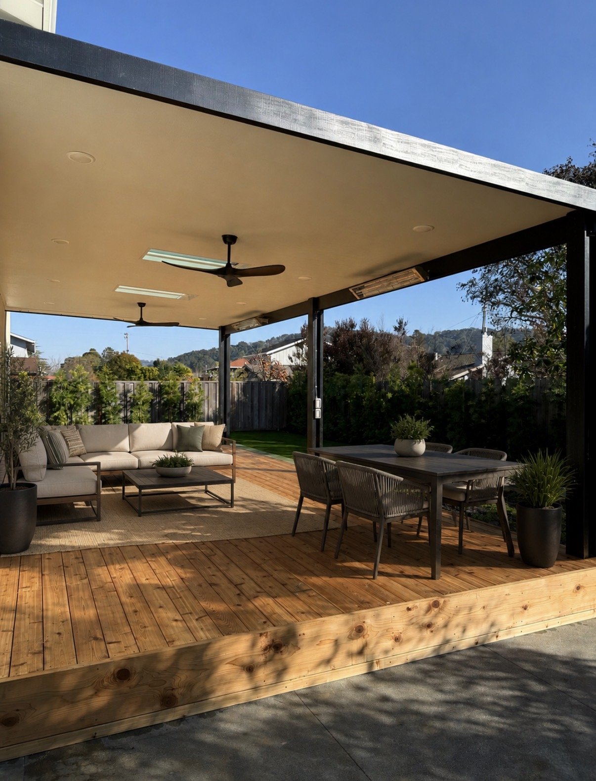 Covered outdoor patio with wooden deck, beige outdoor sofa with pillows, dining table with chairs, potted plants, ceiling fans, and a view of trees and houses under a blue sky.