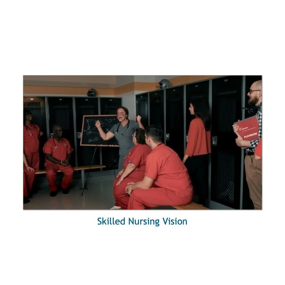 A group of healthcare professionals in a locker room, some wearing red scrubs, listening to a woman in gray scrubs explaining using a chalkboard with diagrams. The setting suggests a team meeting or training session, with the caption "Skilled Nursing
