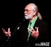 an older white male, with gray beard and gray ponytail, in a black jacket, at a mic performing.