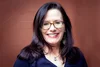 a beautiful smiling white woman with glasses, her blue tinted hair draped on her purple blouse, in front of a brick red background