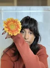 a beautiful black woman holding a flower up by her face, her long brown hair falling over her brown sweater.
