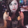a native-americas femme giving the double bird, with a glass of water in front of them and a smirk on their face.