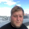 a serious looking white male with facial hair standing on a rooftop, a blue sky above him.