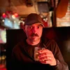 a white man with a full mustache, holding up a glass of whiskey under a dim red tinted light inside a bar.