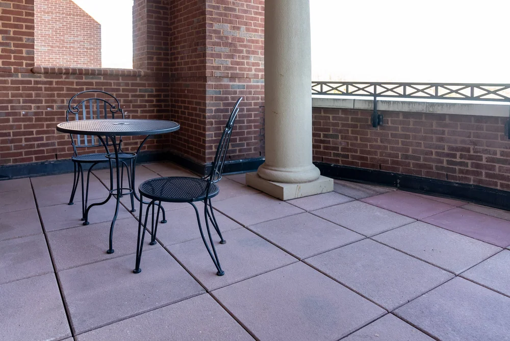 An outdoor terrace with a small black round metal table and two matching black metal chairs, surrounded by brick walls and a stone column.