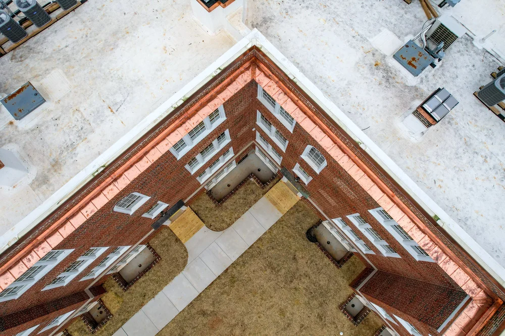 Aerial view of the corner of a brick apartment building with a lawn and concrete walkway, surrounded by a concrete rooftop with HVAC units.