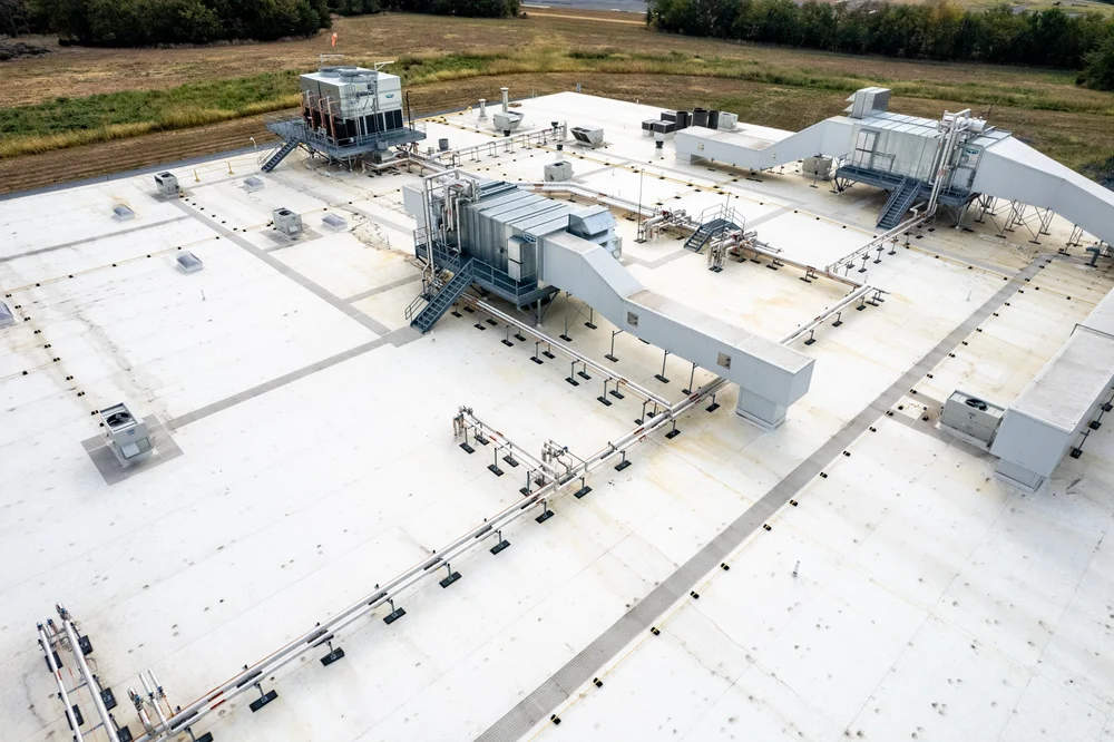 Aerial view of a large white rooftop with industrial HVAC units, ductwork, and ventilation systems, surrounded by open fields and some trees.