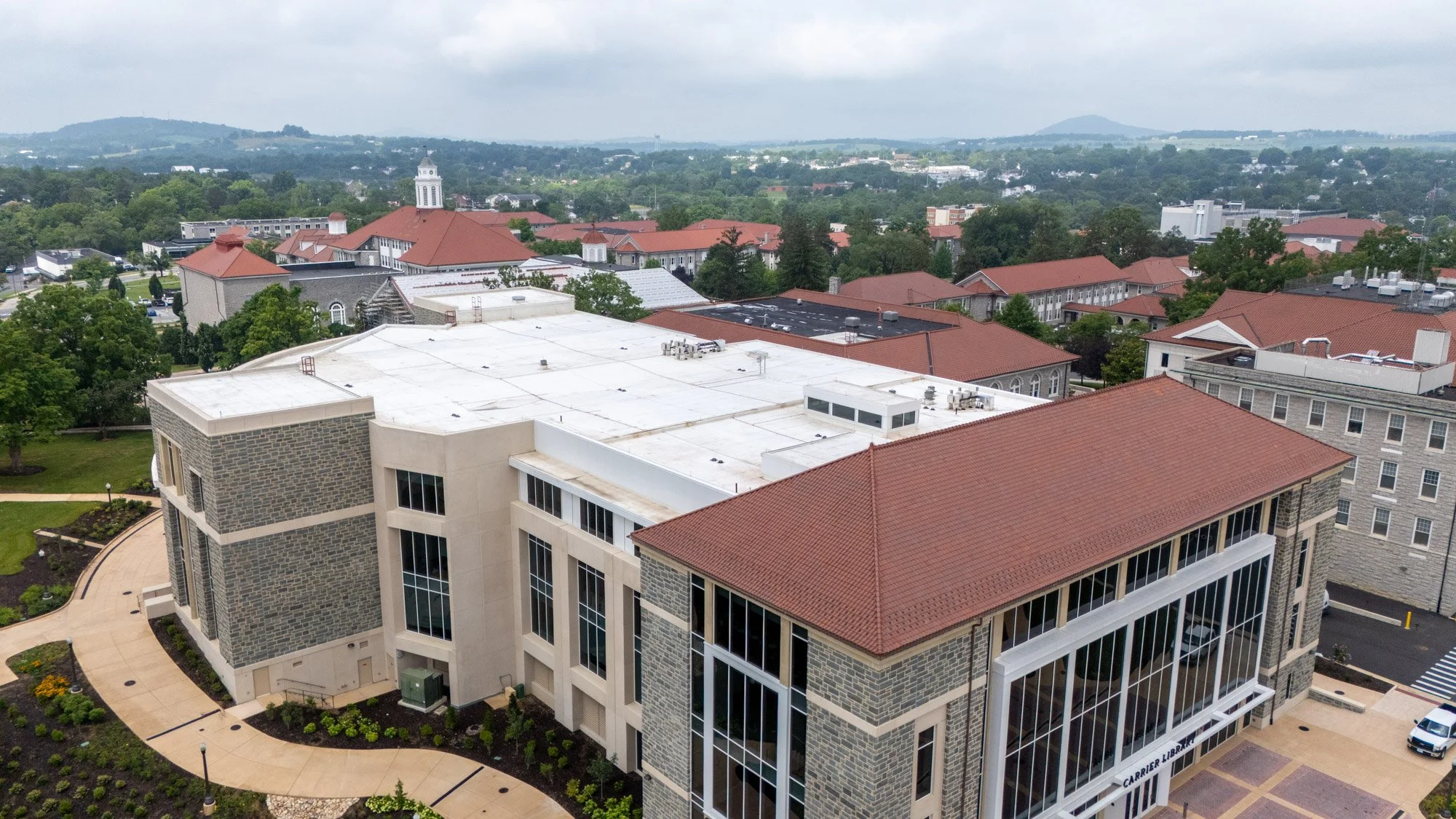 Aerial view of a red clay tile roof on a university building alongside an adjacent white low-slope roof section, with additional campus buildings and hills in the background.