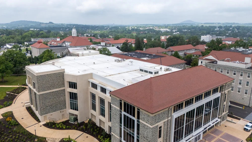 Aerial view of a red clay tile roof on a university building alongside an adjacent white low-slope roof section, with additional campus buildings and hills in the background.
