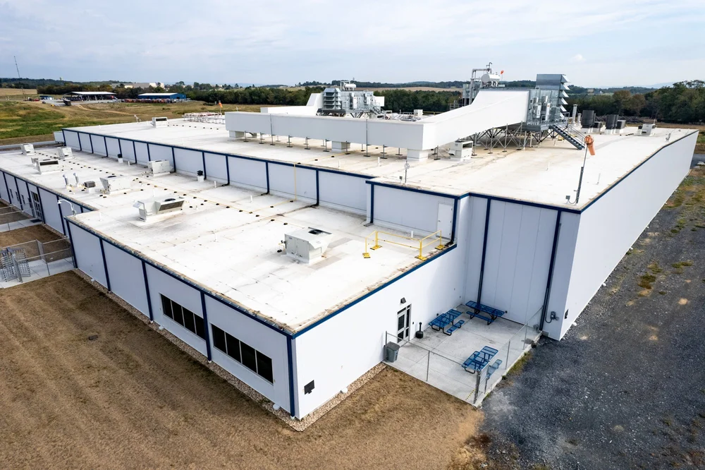 An aerial view of a large industrial building with a white flat roof, ventilation units, and a rooftop stairway, surrounded by a yard with grass and dirt.