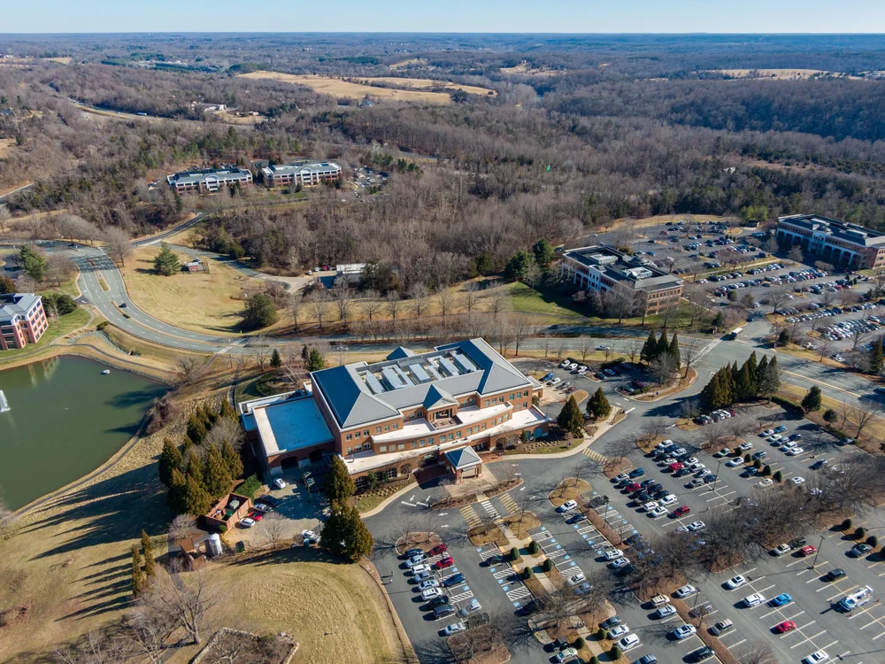 An aerial view of a suburban area with a hospital or large building surrounded by parking lots, roads, a pond, and wooded areas, under clear skies.