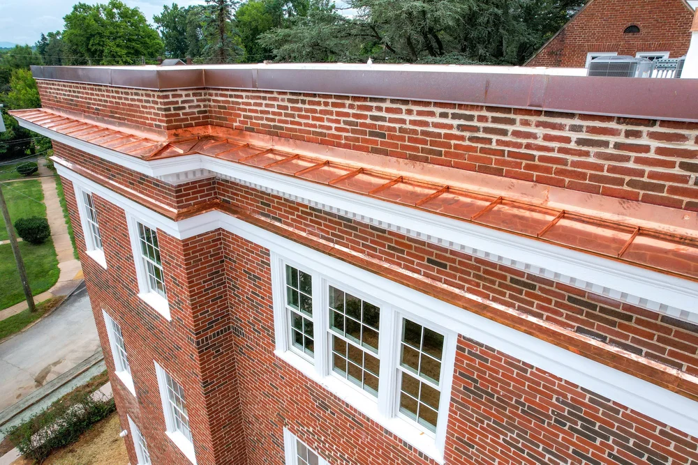 Close-up view of a multi-story brick building with white window trim, a copper roof section, and a brick parapet wall. The building is surrounded by trees and a sidewalk.