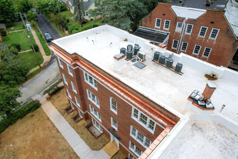 Aerial view of a brick apartment building's flat roof with multiple HVAC units, ventilation pipes, and a chimney, situated in a neighborhood with trees, grass lawns, a sidewalk, and residential houses.