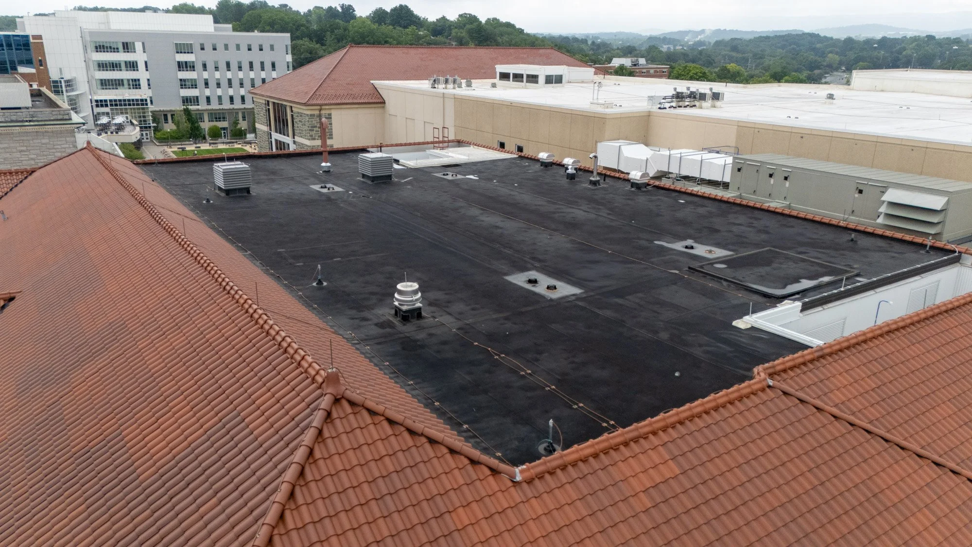 Aerial view of a black EPDM low-slope roof with rooftop vents, a roof hatch, and HVAC equipment, bordered by red clay tile roofing on a campus building.
