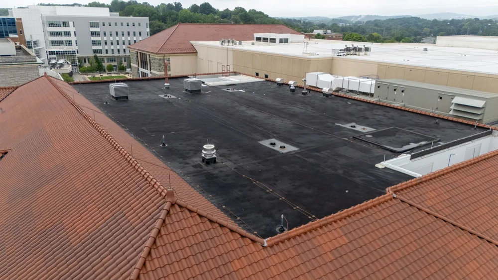 Aerial view of a black EPDM low-slope roof with rooftop vents, a roof hatch, and HVAC equipment, bordered by red clay tile roofing on a campus building.