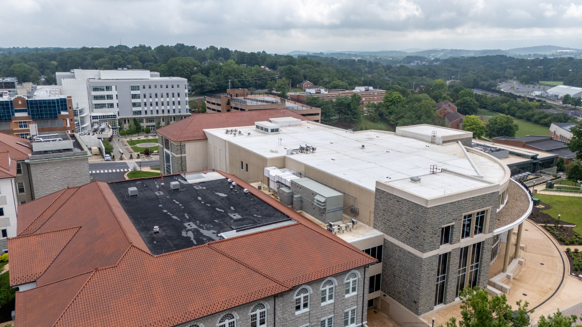 Aerial view of a low-slope commercial roof with white membrane roofing and clay tile roof sections on an education building in Harrisonburg, VA, with rooftop HVAC units and mountain views in the background.