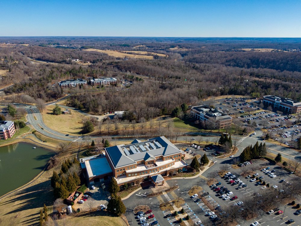 Aerial view of a landscaped campus with parking lots, office buildings, and a pond, surrounded by wooded areas and open fields under a clear blue sky.