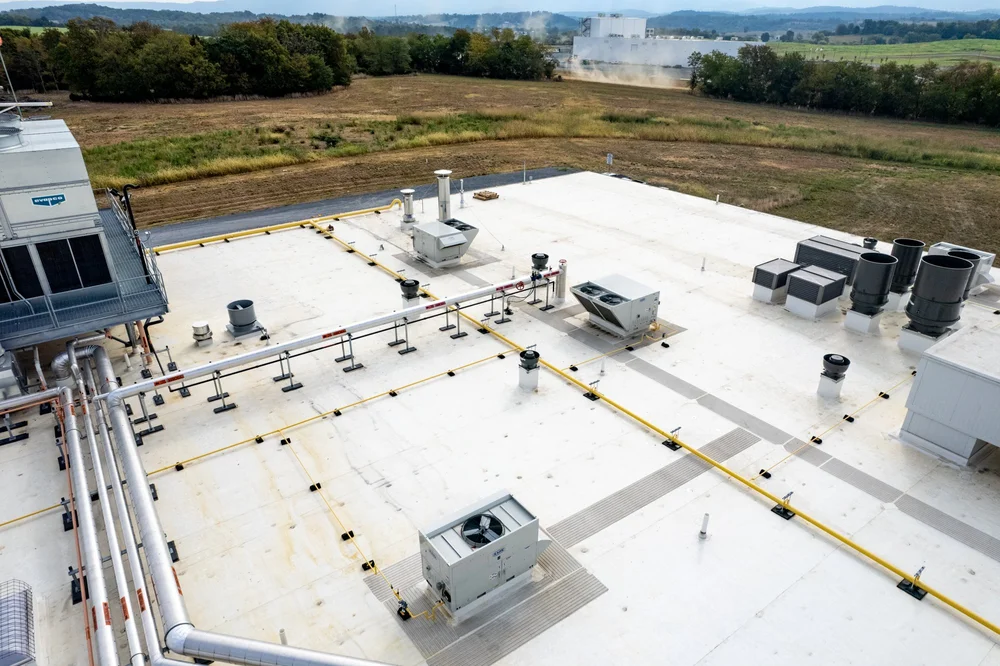 Rooftop of a commercial building with HVAC equipment, vents, and pipes, with a grassy field and industrial area in the background.