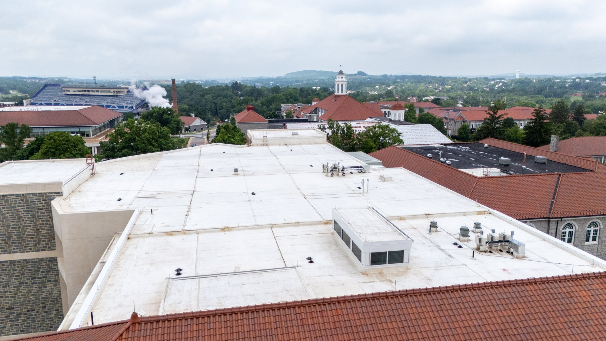 Aerial view of a white low-slope EPDM roof on a campus building, with a clock tower, football stadium, and clay tile roofs visible in the background near Harrisonburg, VA.