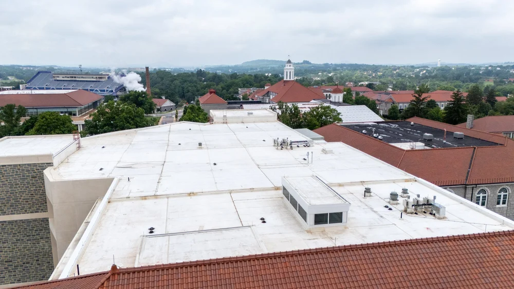 Aerial view of a white low-slope EPDM roof on a campus building, with a clock tower, football stadium, and clay tile roofs visible in the background near Harrisonburg, VA.