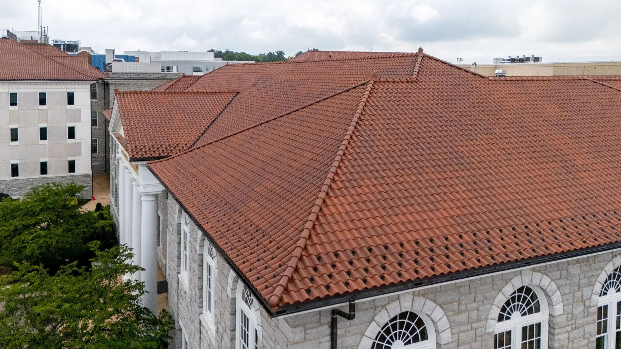  Aerial view of a red clay tile roof with dark hip caps and dentil-style trim on a stone campus building, showing classic architectural roofing details.