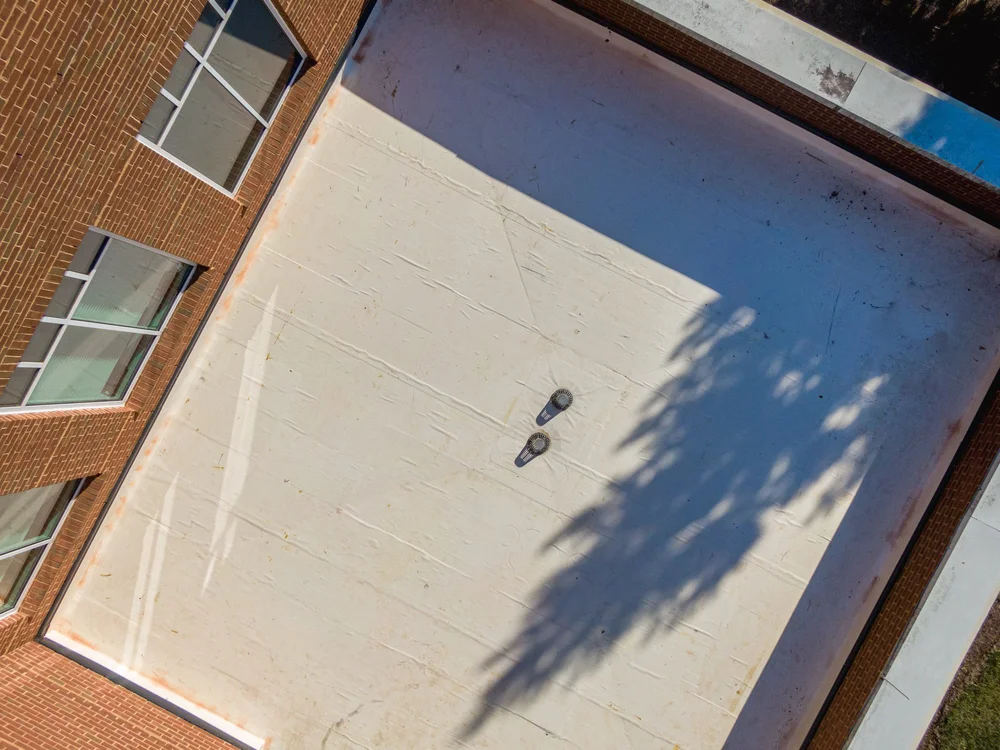 Aerial view of a rooftop with white membrane, brick building walls on three sides, two rooftop vents, and shadows of trees.