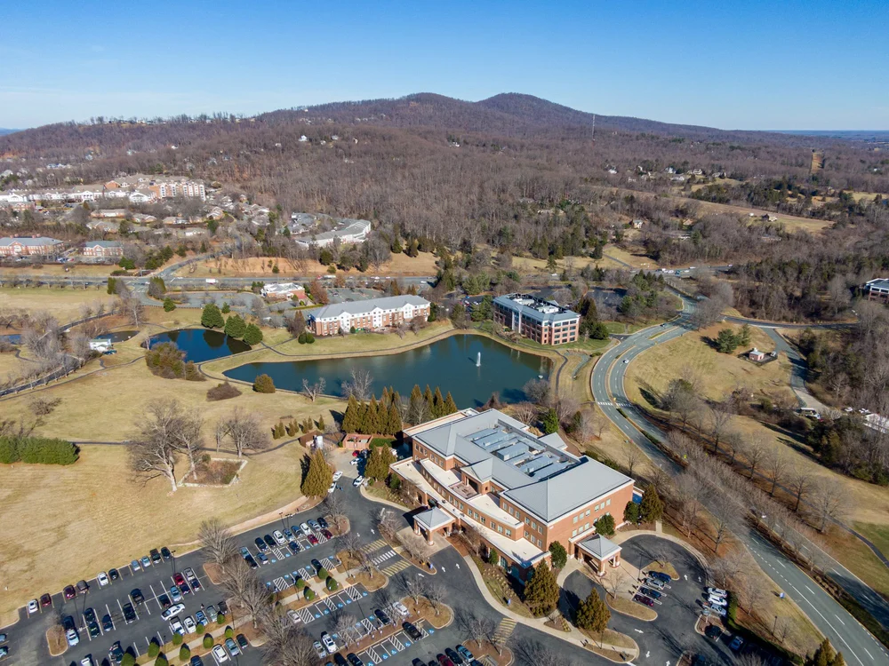 Aerial view of a campus with a lake, several buildings, roads, trees, and hills in the background, during winter with leafless trees.
