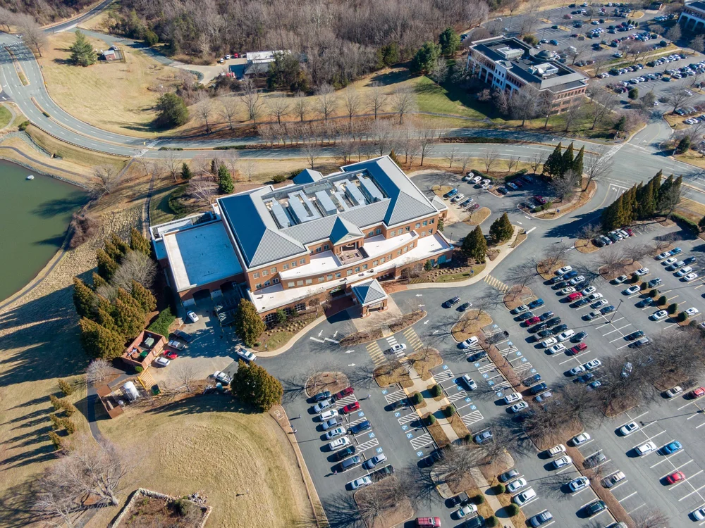 Aerial view of a large building with a parking lot, surrounded by landscaped areas, trees, and a pond, with additional buildings and roads nearby.