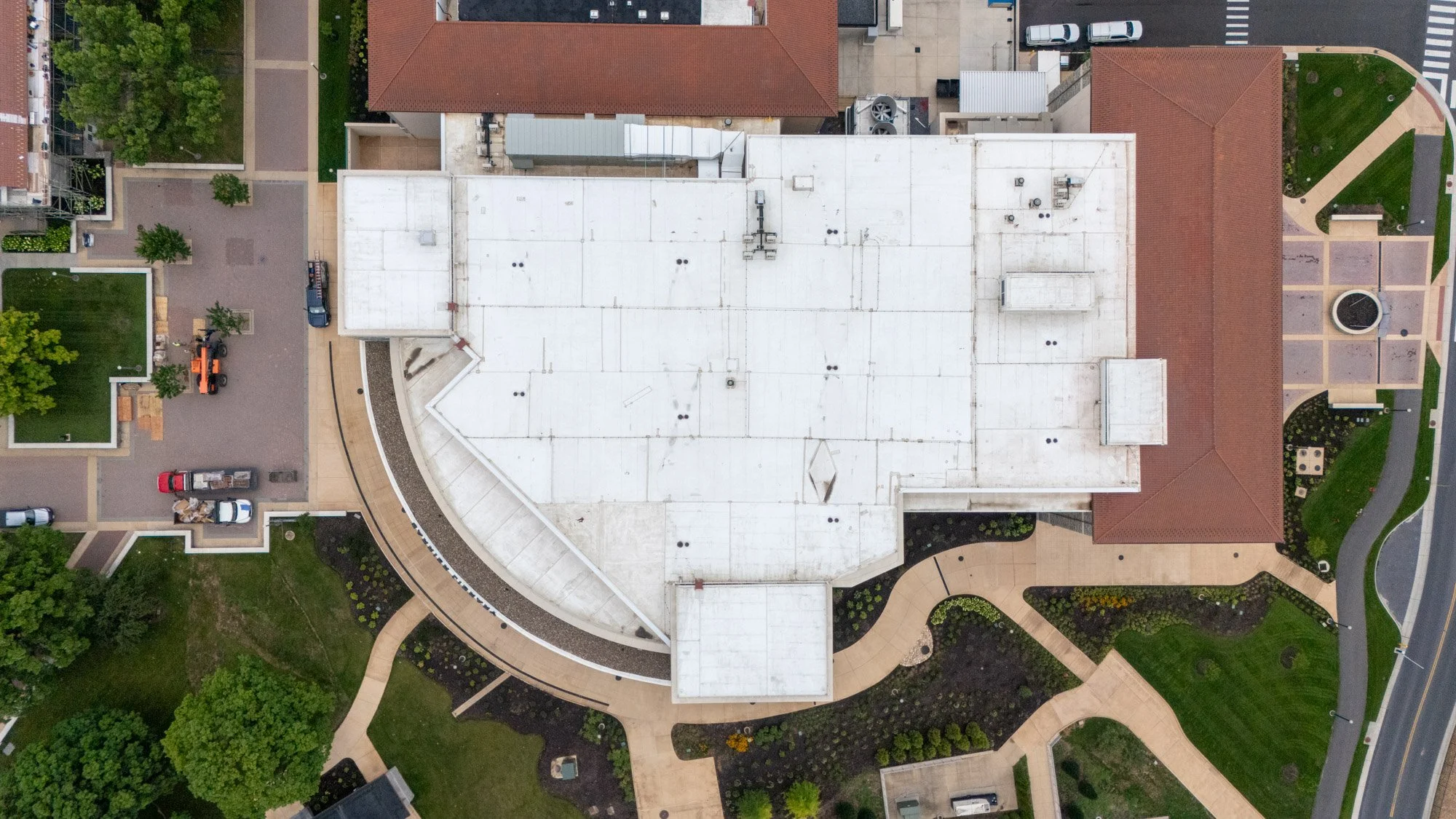 Top-down aerial view of a curved white commercial roof adjoining a clay tile roof, surrounded by landscaped walkways and courtyard space.