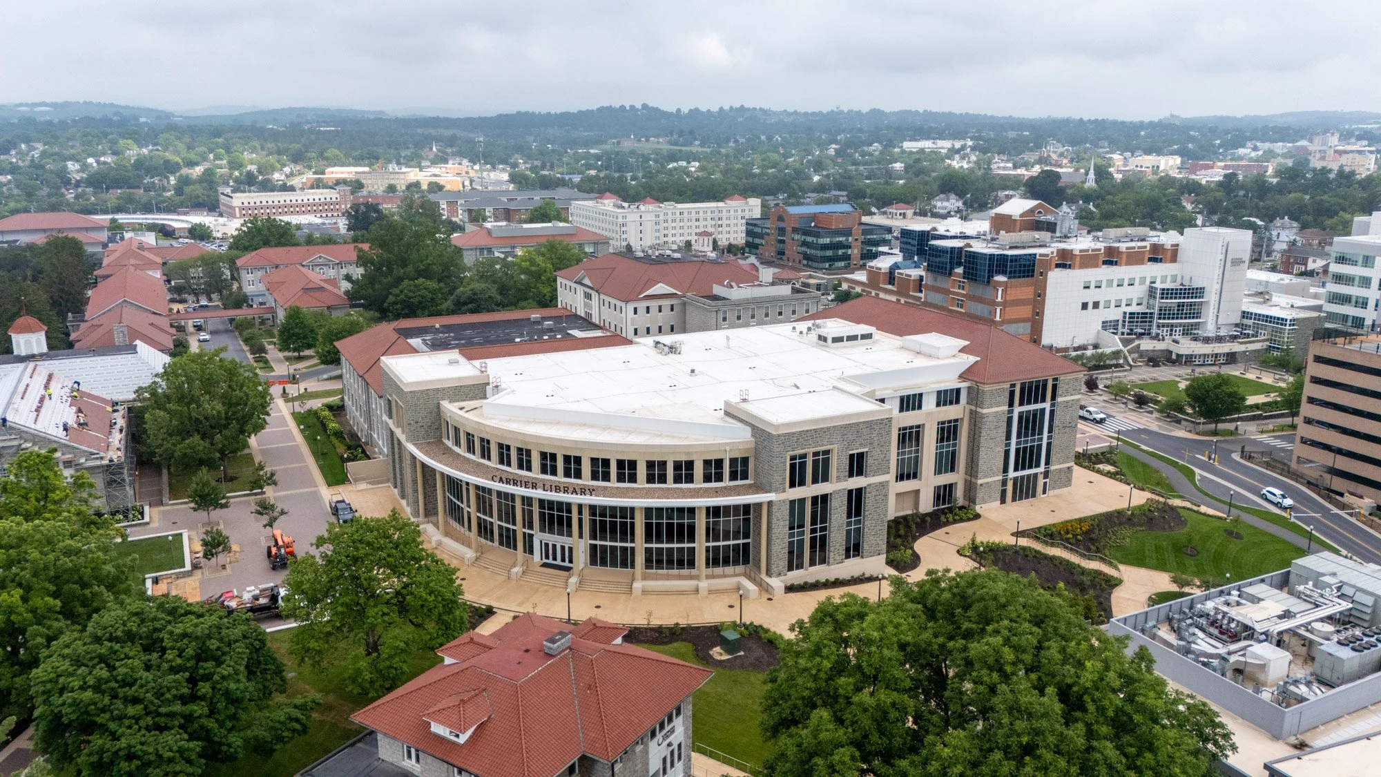 Aerial view of Carrier Library at James Madison University, showing its white low-slope roof and curved glass facade, with the surrounding campus and downtown Harrisonburg skyline in the distance.