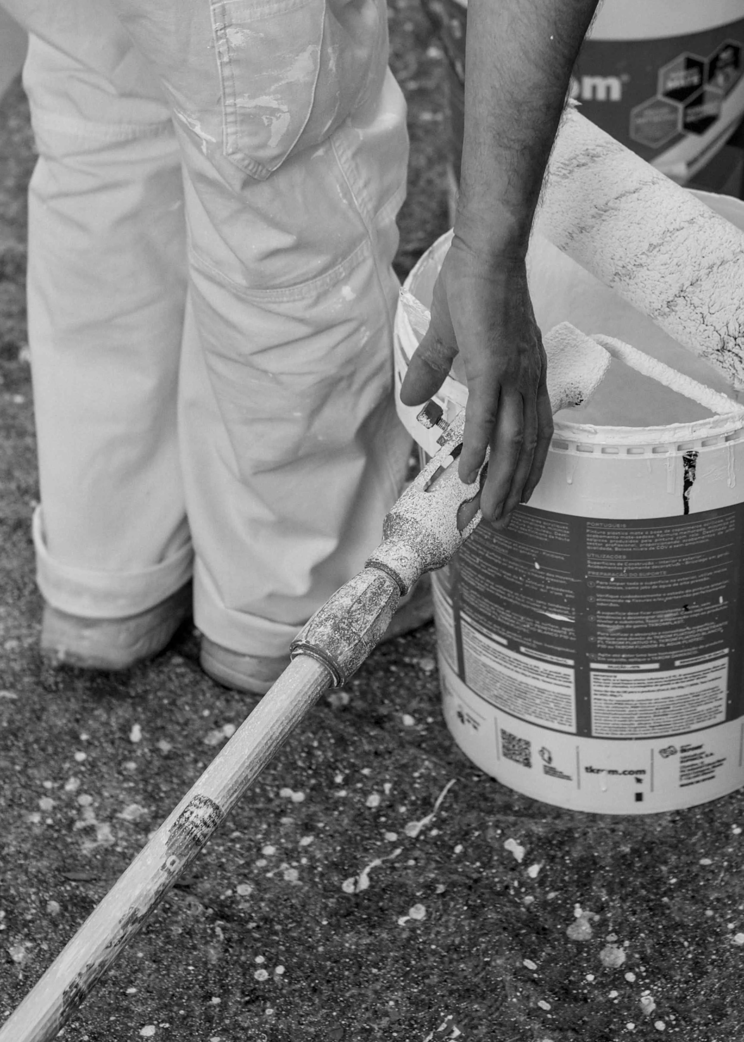 Close-up of paint-splattered roller and bucket beside painter’s legs.