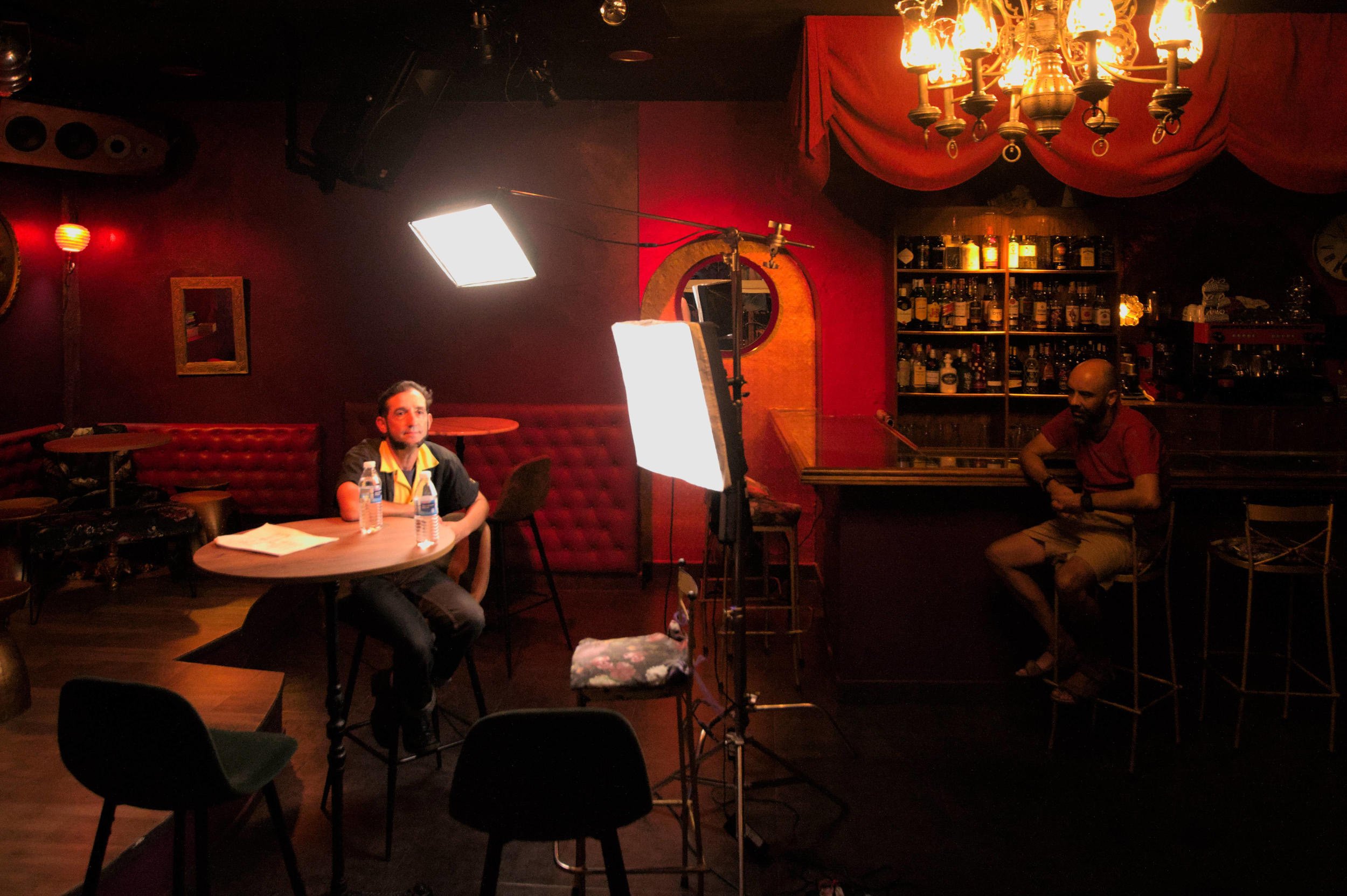 Actor waiting at a bar table under two bright softboxes in a red-lit bar.