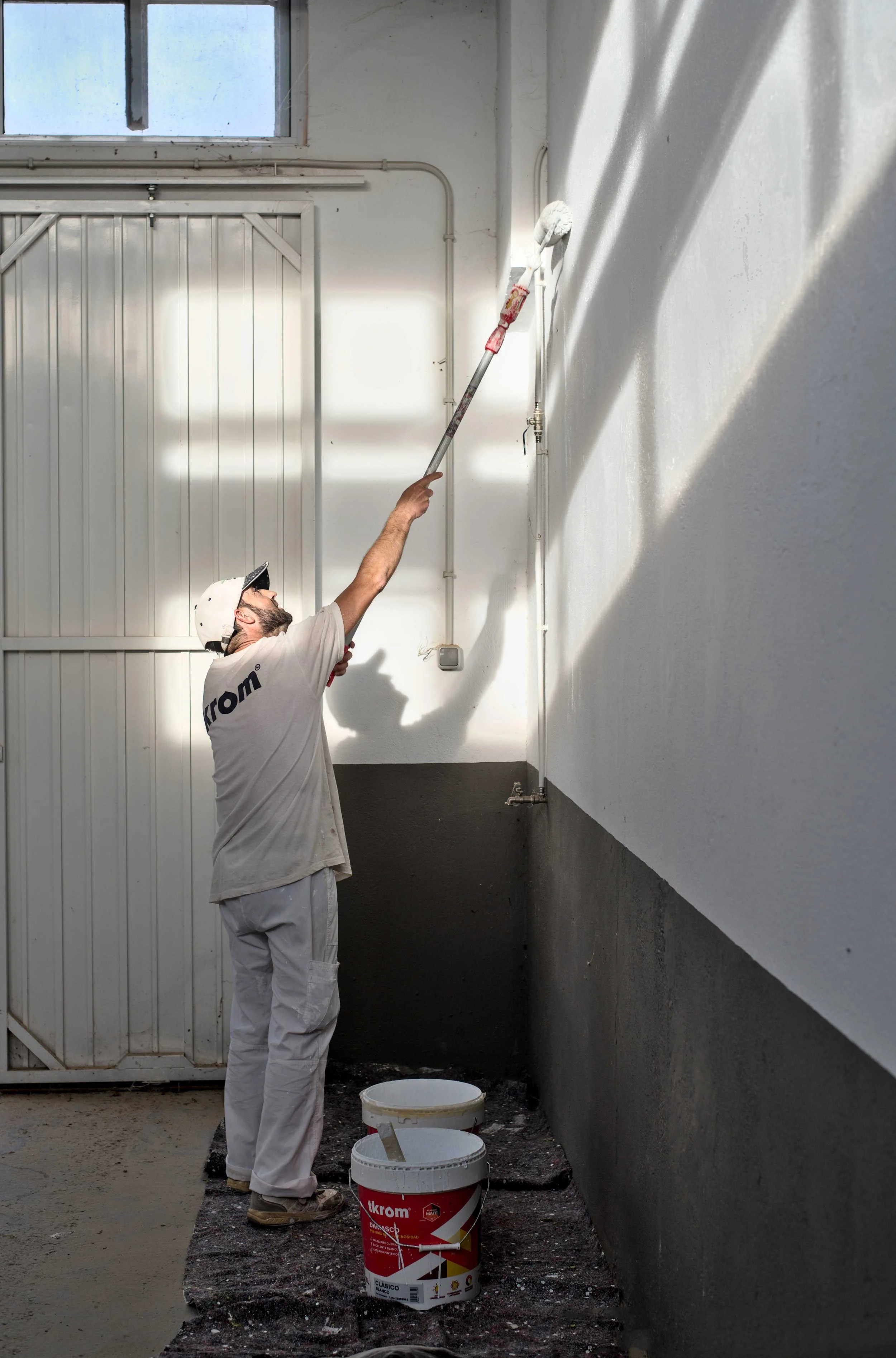 Painter reaching up with a roller to paint a high interior wall in strong light.