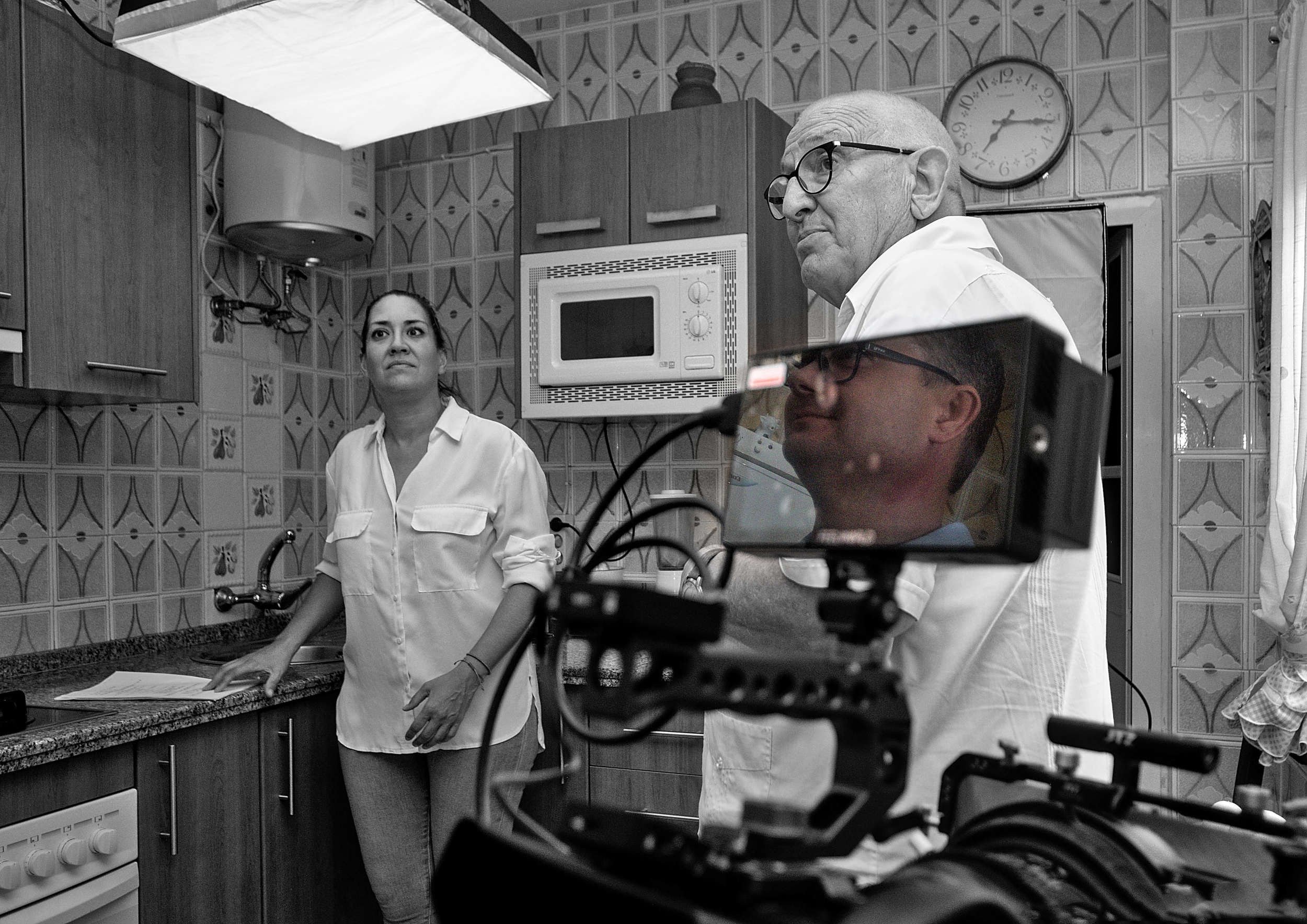 Actress and older actor in a tiled kitchen with the DP reflected in the camera monitor.