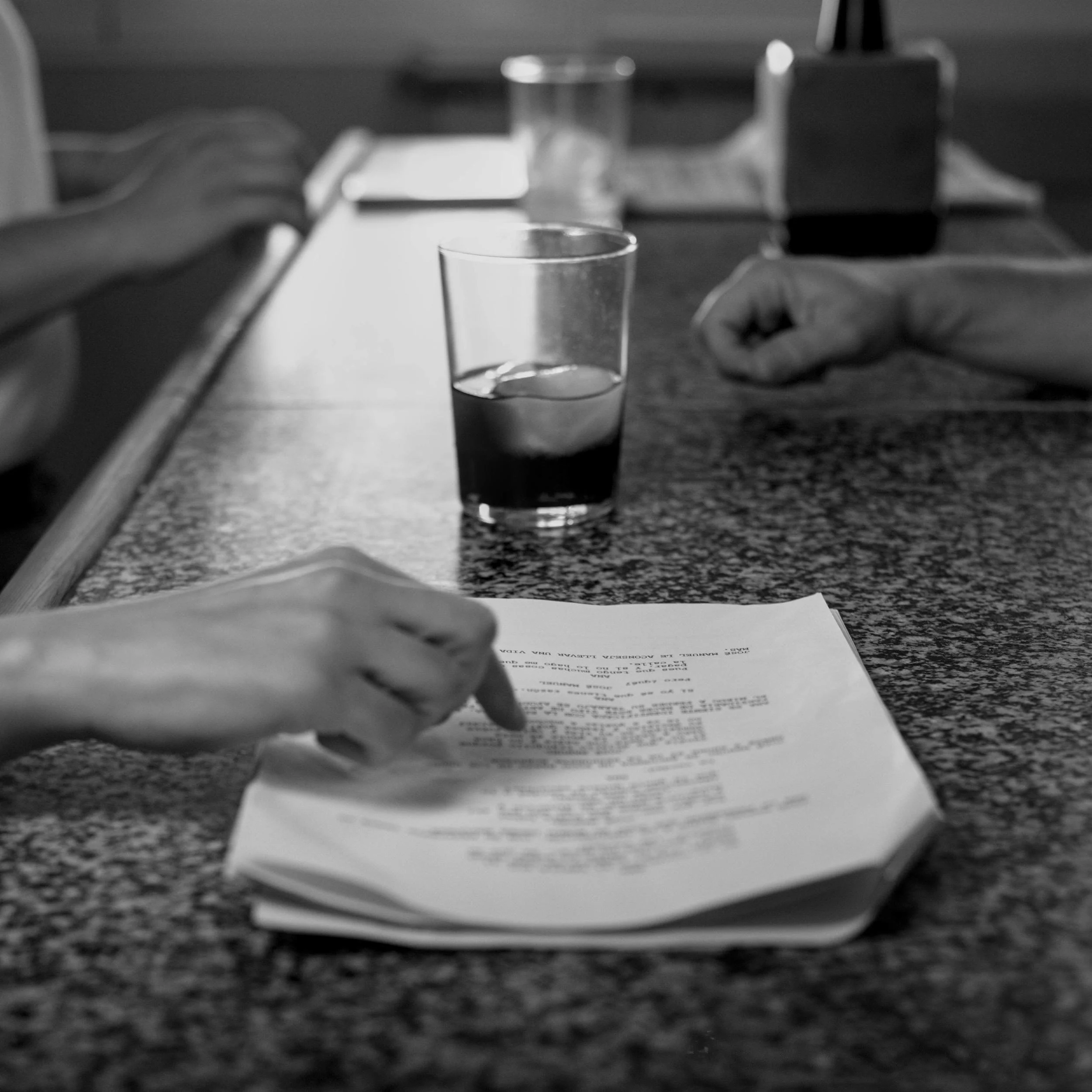 Close-up of hands pointing to a film script on a bar counter beside a drink.