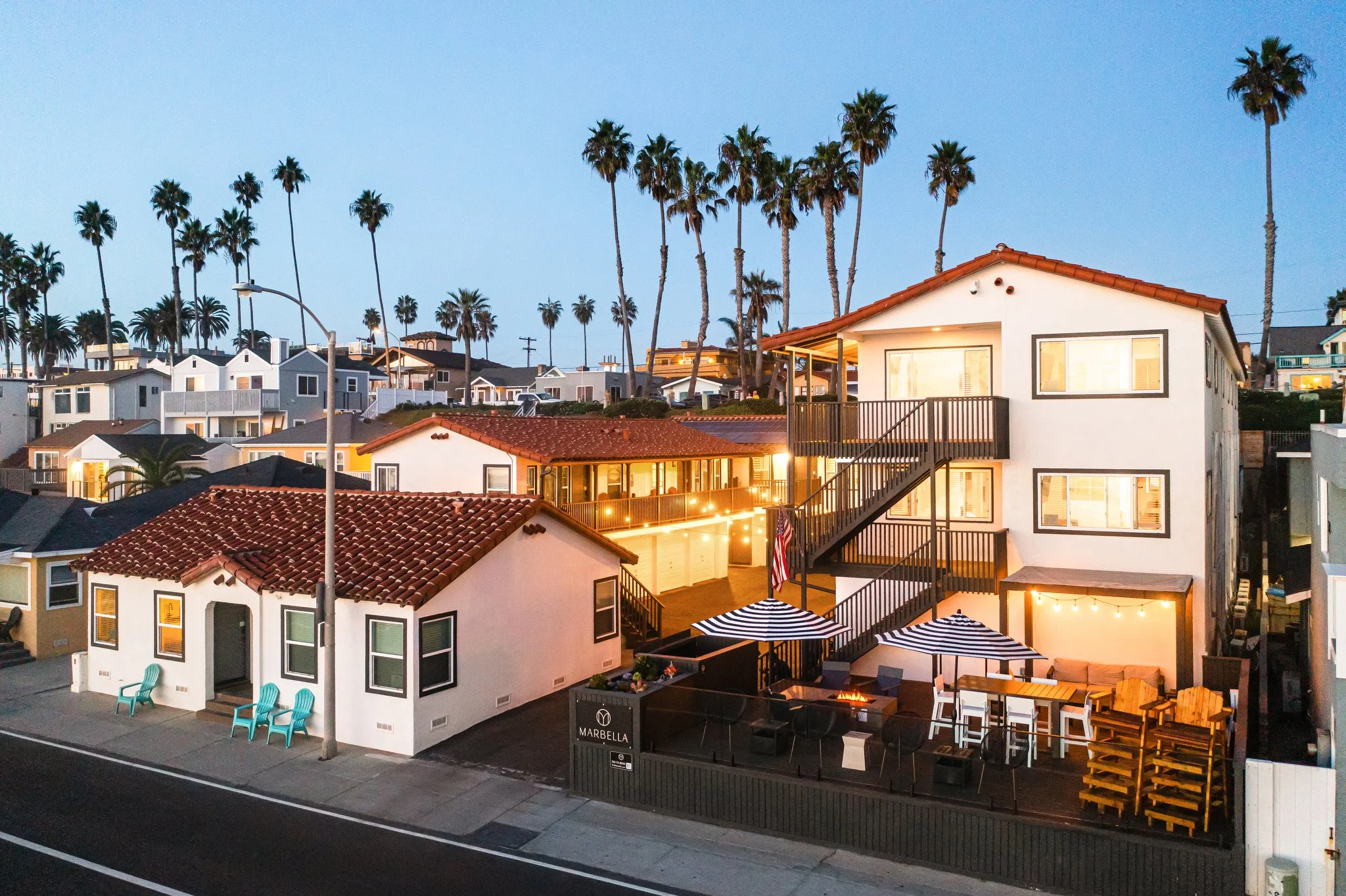 1-Bedroom Vacation Rentals in Oceanside, CA - Marbella - Courtyard Patio Features Warm String Lights, Multiple Balconies, Palm Trees, And Umbrellas Along A Street At Dusk.