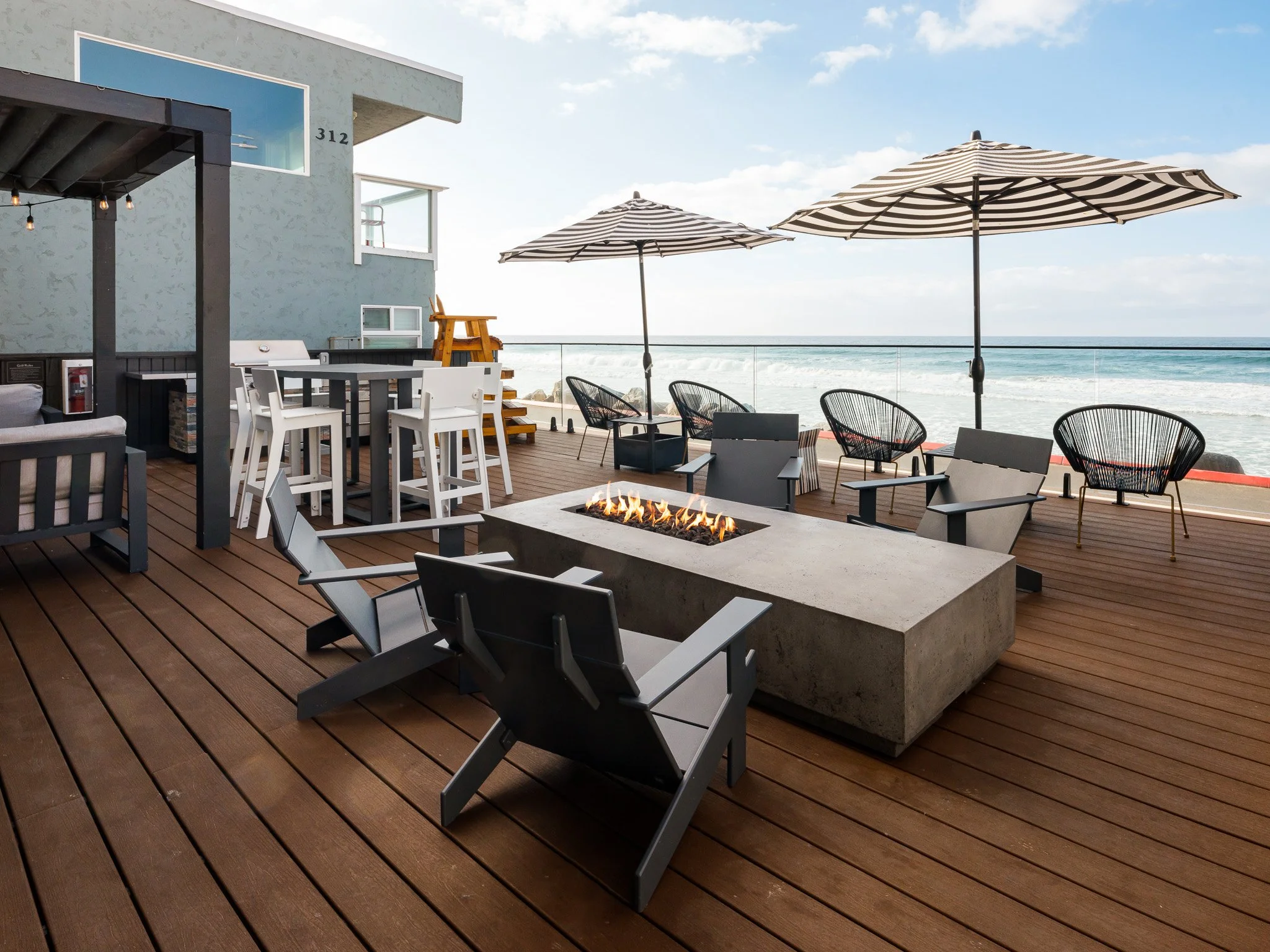 Oceanside, CA, Short Term Leases - Marbella - A Rooftop Fire Pit Area Features A Rectangular Concrete Fire Table With Flames, Surrounding Lounge Chairs, And Ocean Views Under Striped Umbrellas.