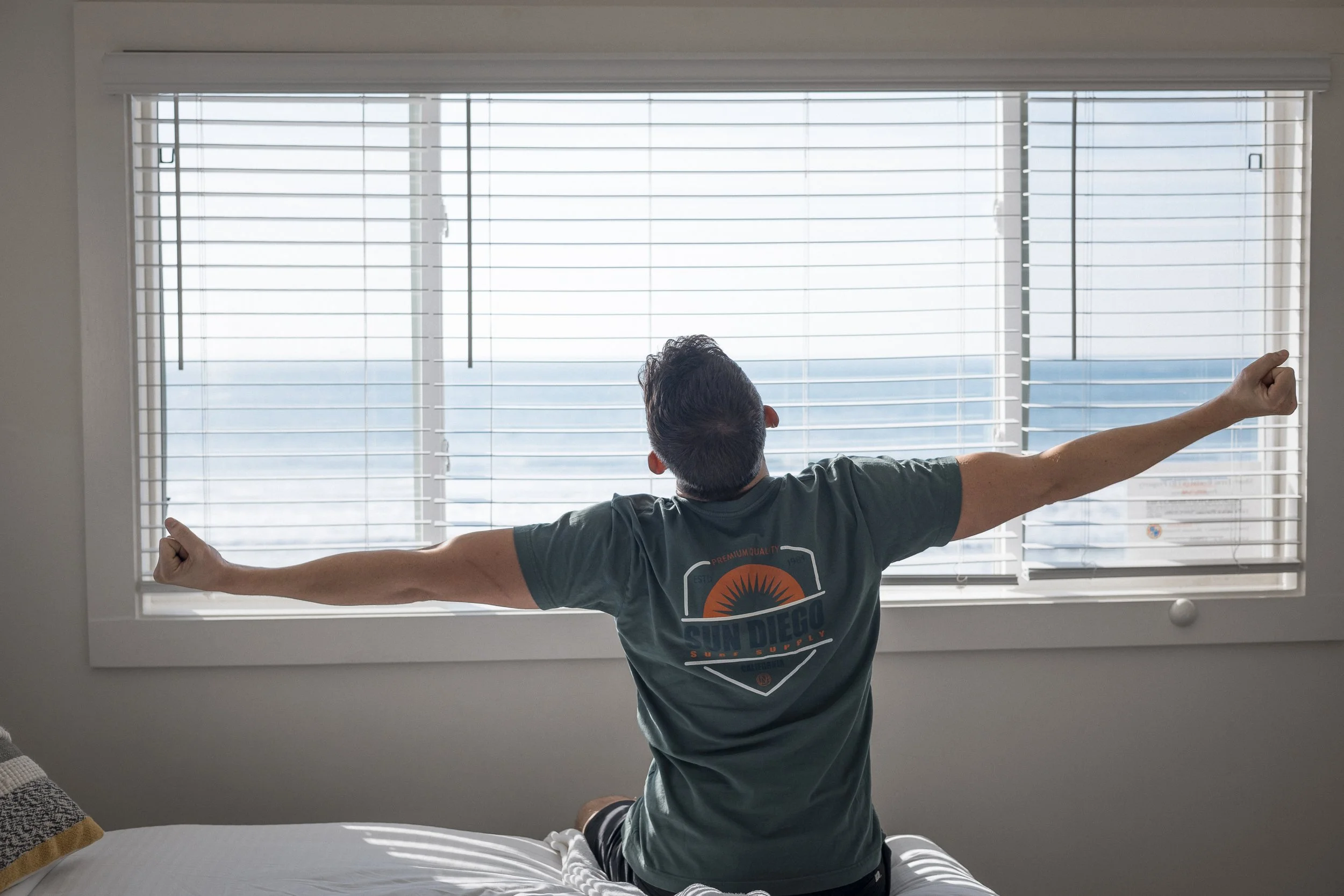 Two-Bedroom Vacation Rentals in Oceanside, CA - Marbella - A Bedroom With A Person Sitting On A Bed Stretching Their Arms Toward A Large Window With Blinds, Looking Out At A Bright Ocean View.