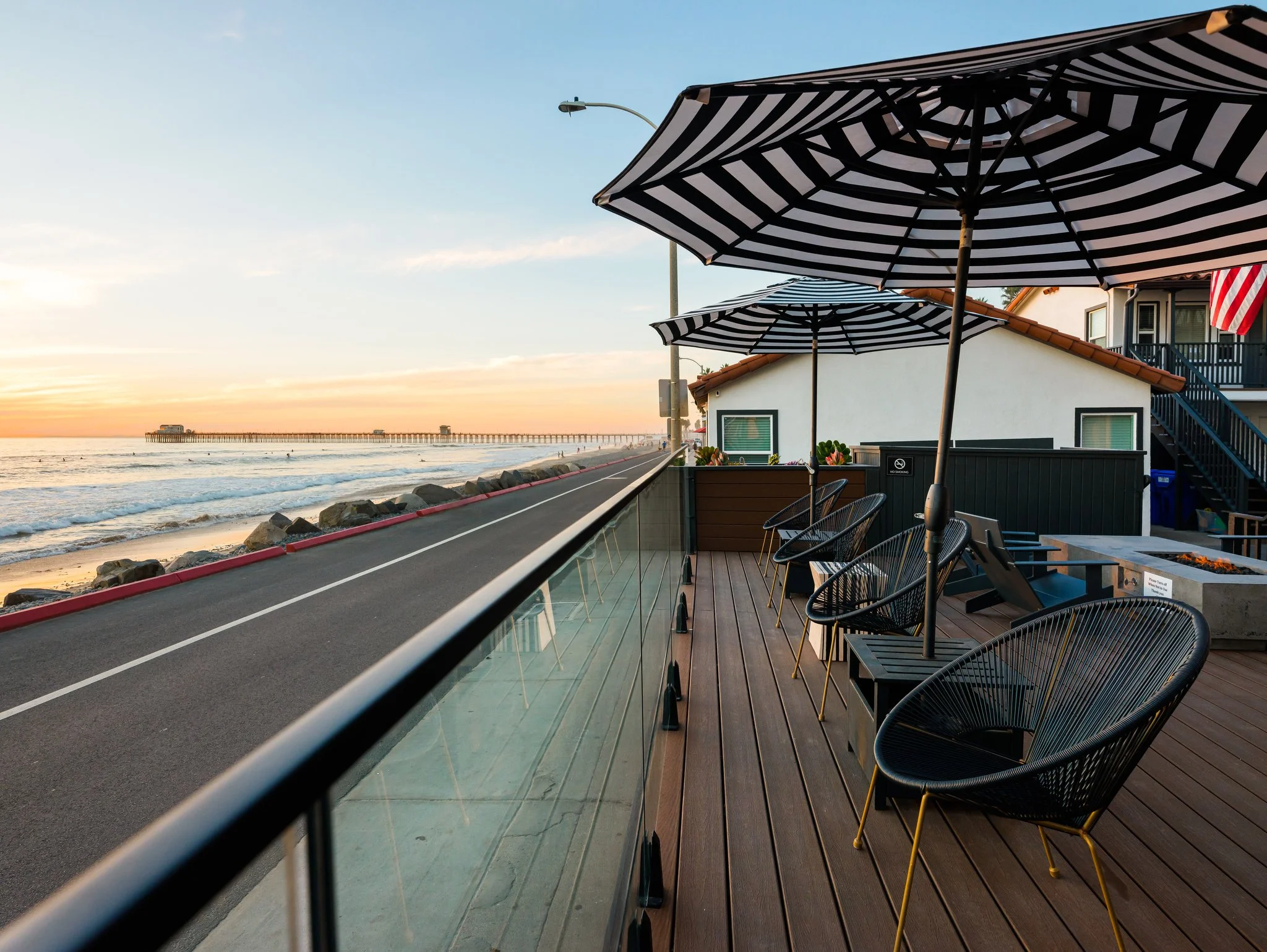 Short Term Leases in Oceanside, CA - Marbella - An Outdoor Patio Features Umbrella Shaded Seating Along A Glass Railing With A Coastal Road And Ocean View At Sunset In The Background.