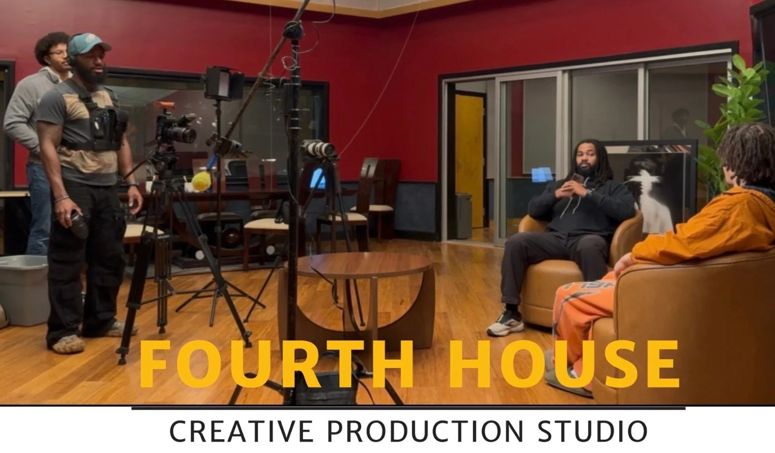Inside a creative production studio with two men sitting on chairs having a conversation, while three crew members operate filming equipment in front of them. The studio has red and black walls, wooden flooring, and large windows.