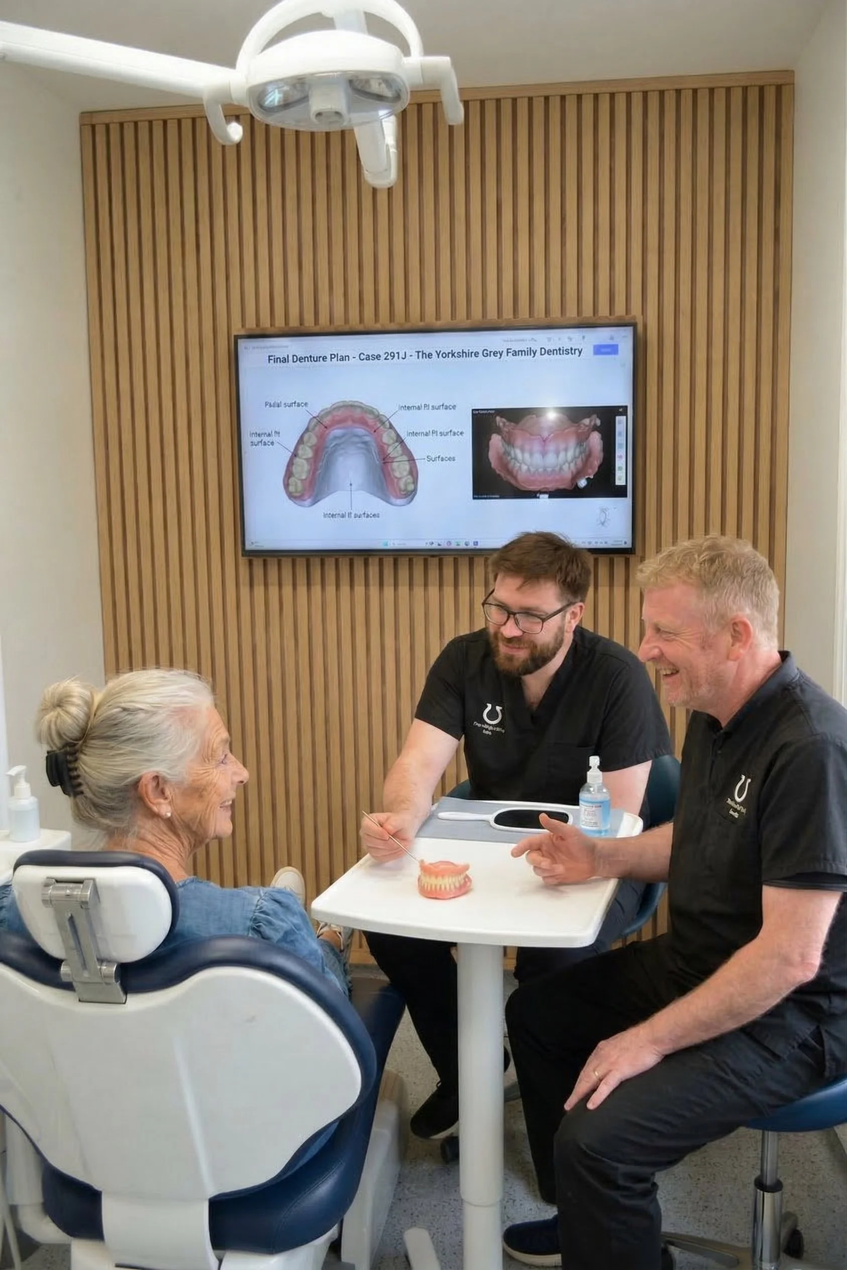 A dentist and an assistant talking to an elderly woman patient in a dental office, with dental diagrams and a 3D dental model on the table.