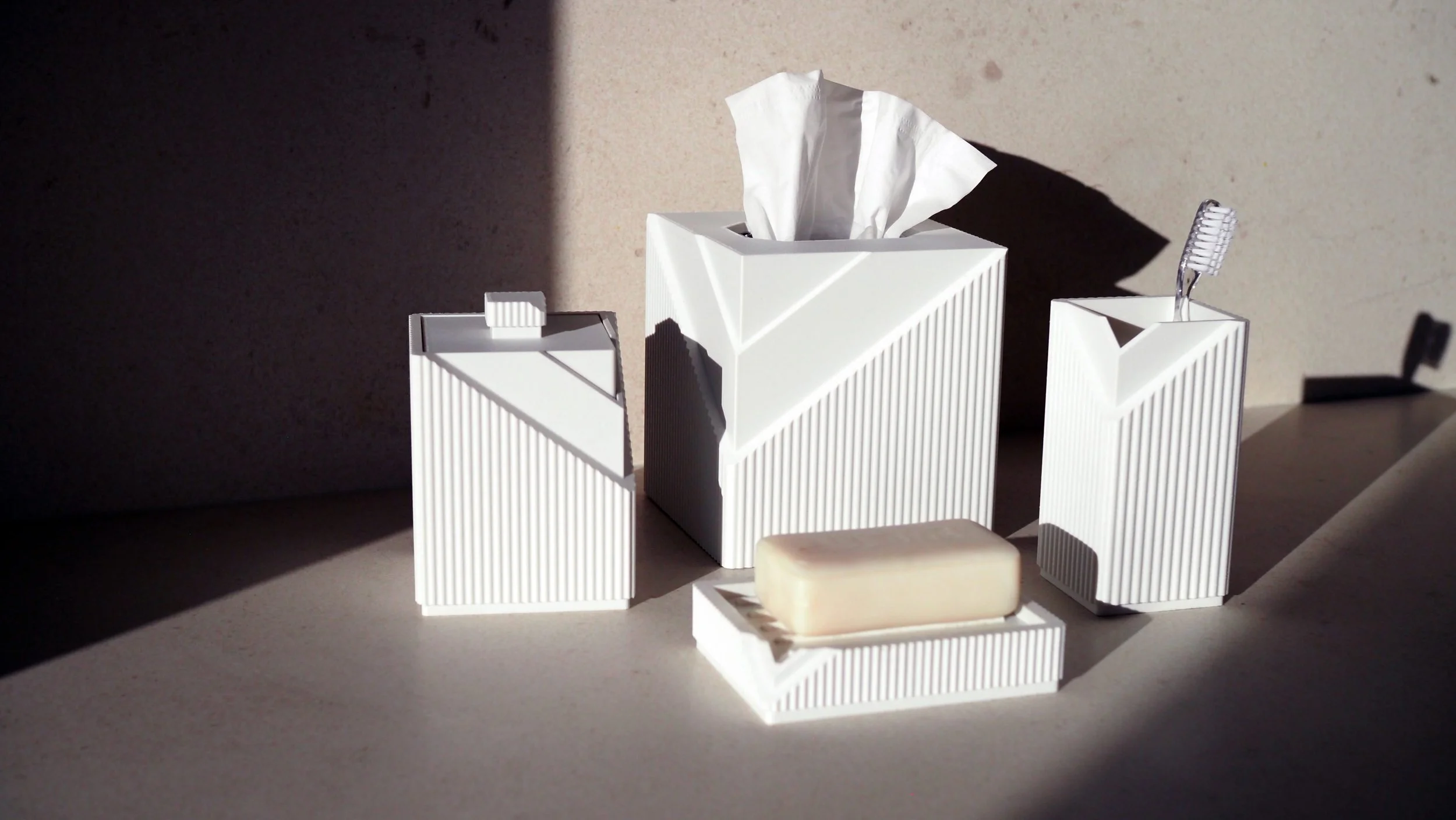 Set of white bathroom accessories including a tissue box with tissue, a soap dish, and a toothbrush holder with a toothbrush, placed on a surface with a shadowed background.