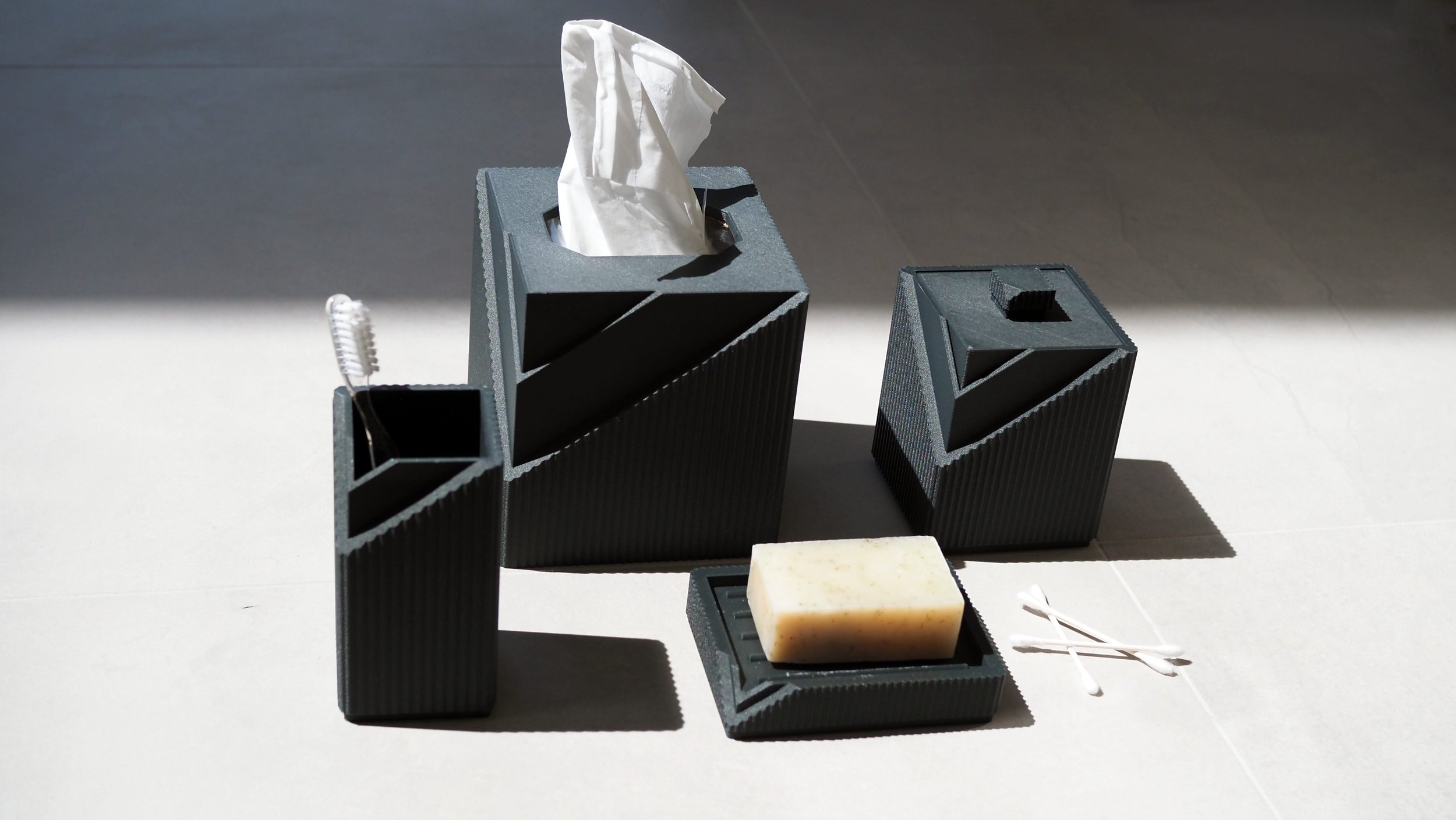 Black textured tissue box with a tissue protruding from the top, a smaller black container with a bar of soap inside, and cotton swabs on a surface with shadows and a neutral background.