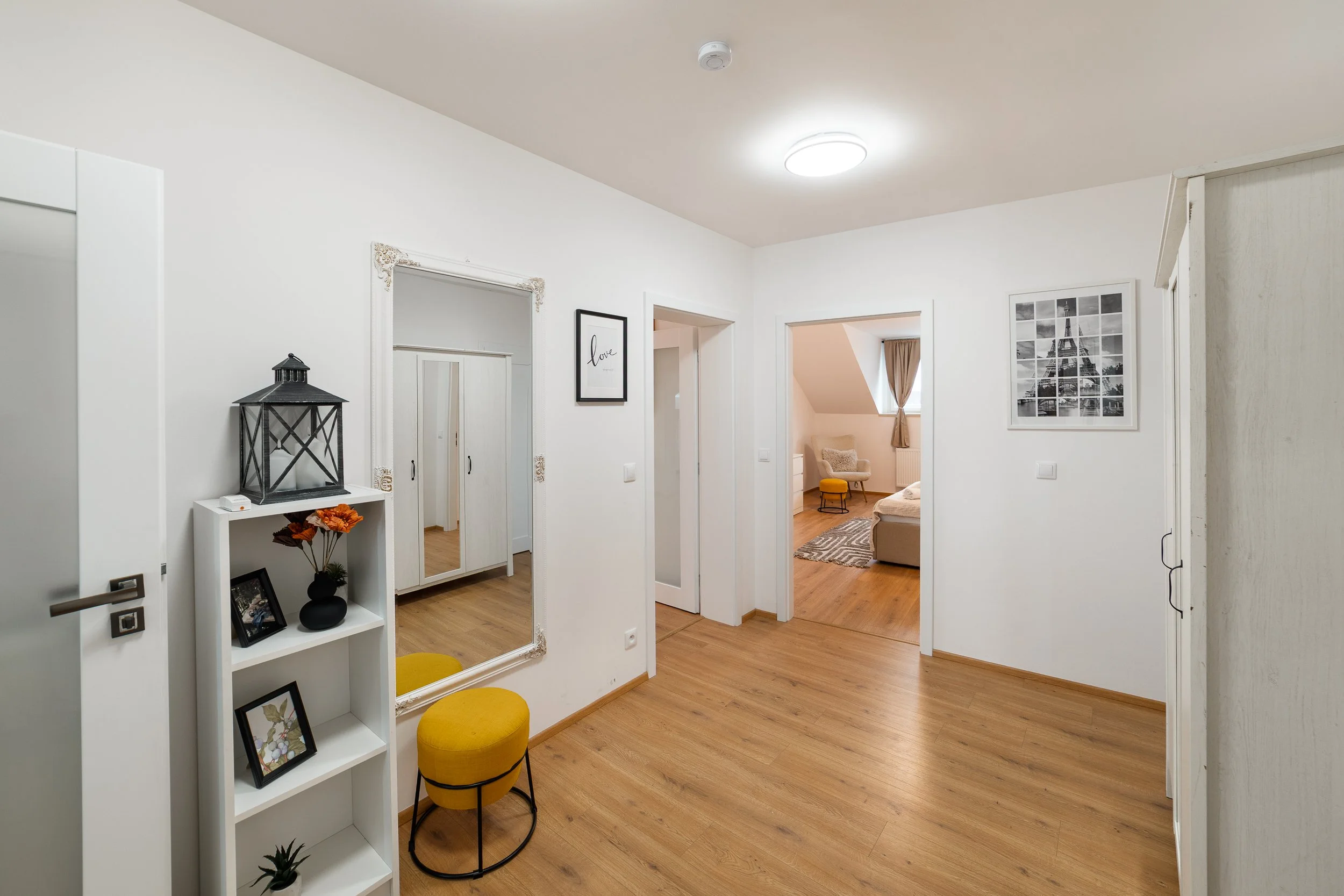 A bright living room with wooden flooring, white walls, and multiple doorways leading to other rooms. Decor includes framed pictures, a black lantern, a small plant, two yellow stools, and a mirror reflecting part of the room. A grey and white patter